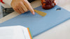 A person holds a jar of California honey and uses a gold-colored butter knife on a blue cutting board, with an open book nearby.