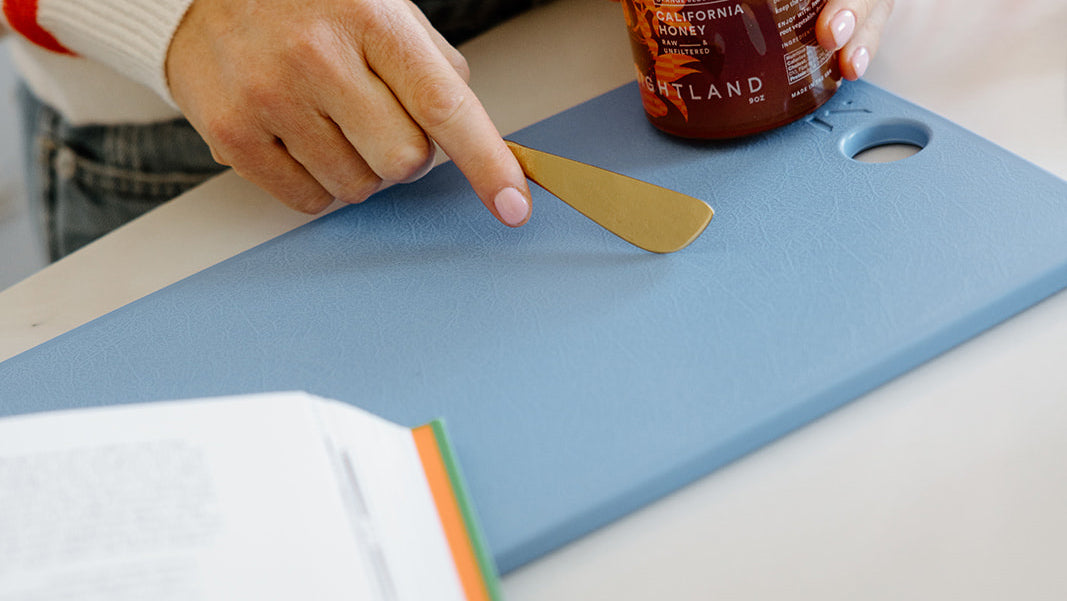 A person holds a jar of California honey and uses a gold-colored butter knife on a blue cutting board, with an open book nearby.