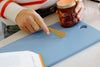 A person holds a gold butter knife above a blue cutting board, with a jar of honey and an open book visible nearby.