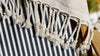 Close-up of a cream-colored fringed blanket draped over a black and white striped fabric on a wooden chair.