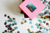 A partially completed jigsaw puzzle is scattered on a white surface next to an open pink puzzle box featuring an image of a floral still life.