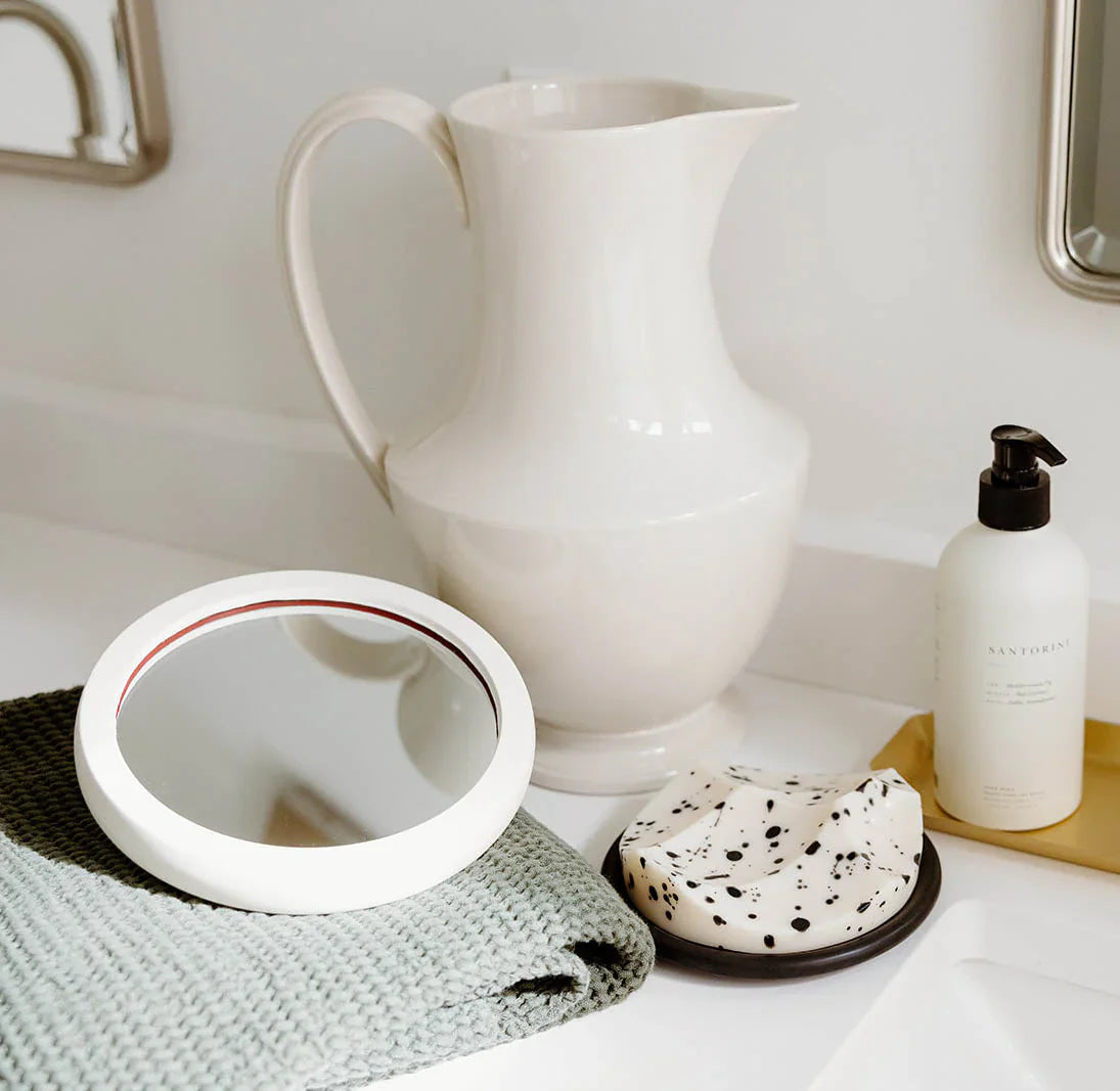 A white pitcher, round mirror on a gray towel, speckled soap on a dish, and a pump bottle are arranged on a bathroom countertop.