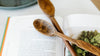 Two wooden spoons rest on an open cookbook displaying text and a photo of leafy greens.