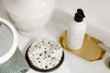 A black and white speckled soap dish, a white bottle with a pump on a gold tray, and a mirror sit on a bathroom counter.