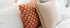 Two decorative pillows rest on a bed; one pillow is beige with red piping, and the other has an orange cover with a white animal pattern. White bedding is visible in the background.