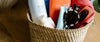 A woven basket containing sunscreen bottles, a pair of sunglasses, a rolled-up red cloth, and a fabric pouch on a wooden surface.