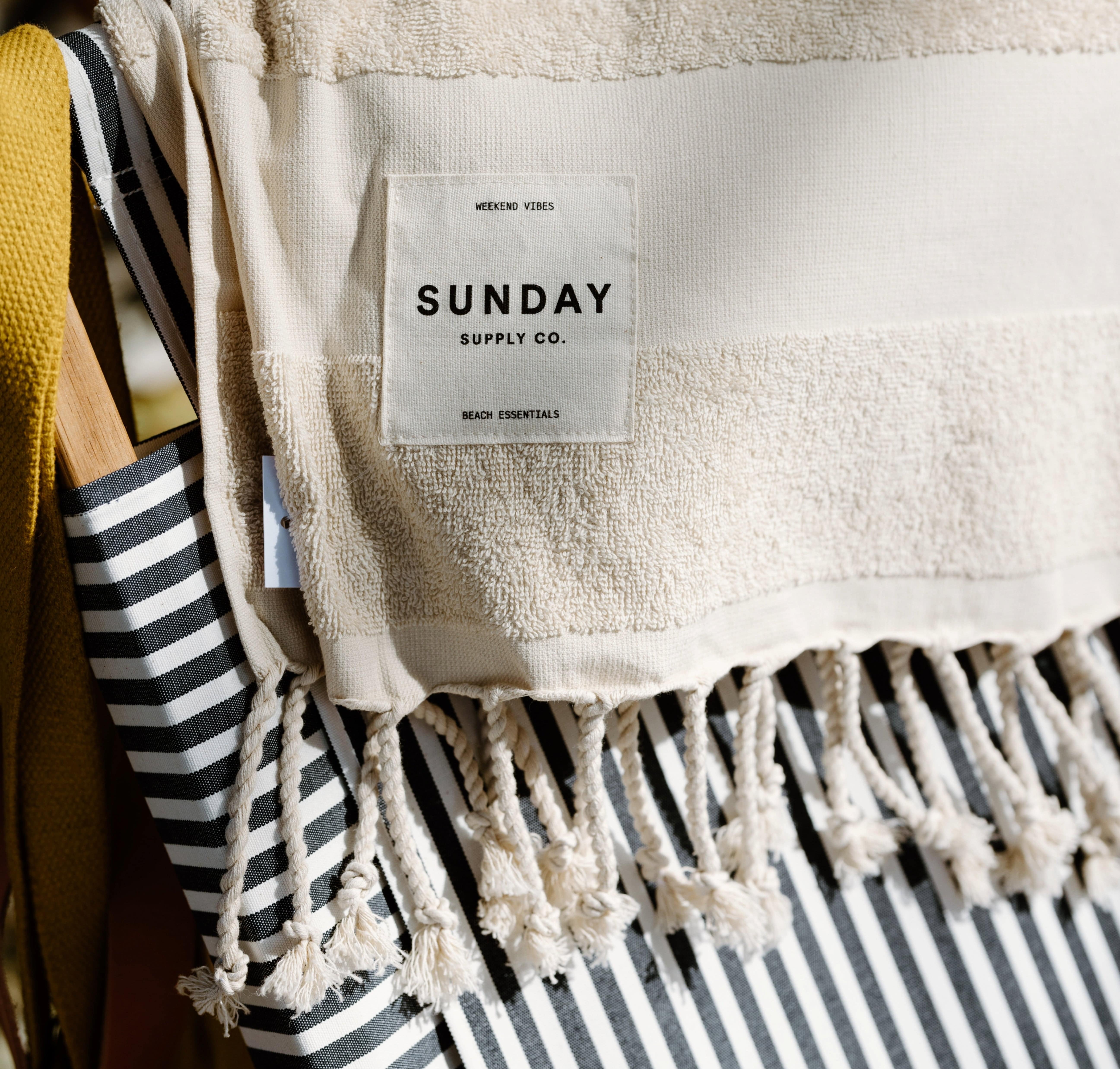 A beige towel with fringe and a Sunday Supply Co. label is draped over a black and white striped bag.