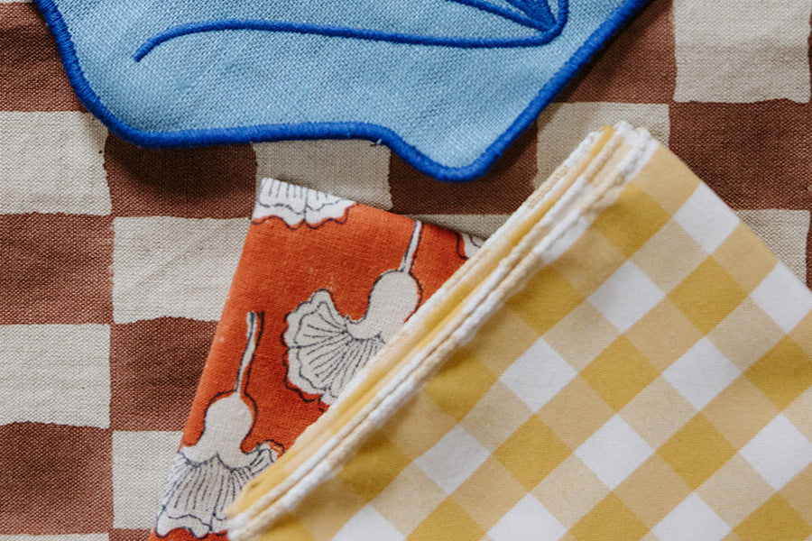 A close-up of three patterned fabrics: blue and brown shapes, orange with white floral design, and yellow gingham checks, layered together.