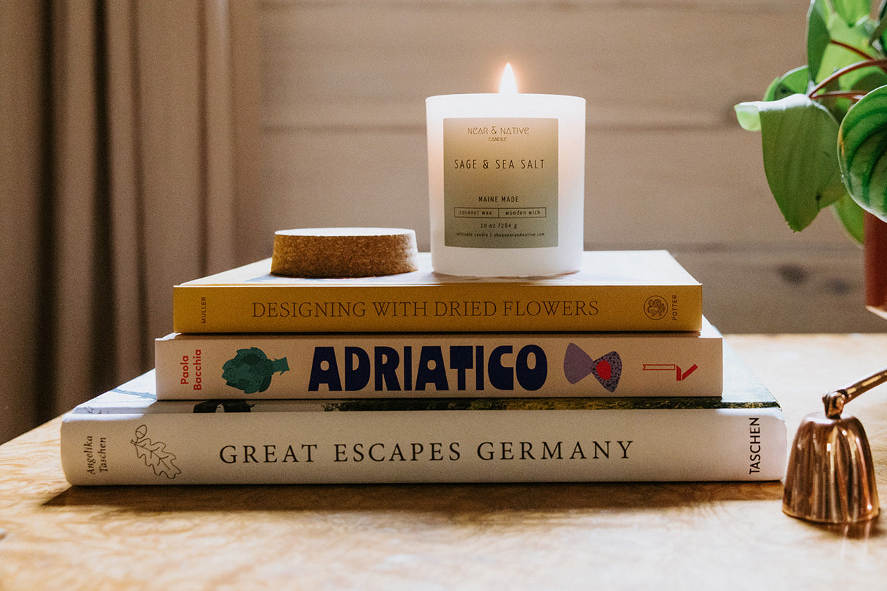A lit candle and a cork lid rest on top of a stack of three books: Designing With Dried Flowers, Adriatico, and Great Escapes Germany, on a wooden table.
