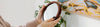 A person holding a round mirror with a brown frame near a wall with a colorful painting and a green plant in the background.