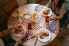 Two people sit at a round table set with croissants, drinks, candles, and decorative napkins on a checkered tablecloth.