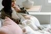 A woman sits on a couch, wrapped in a fluffy white blanket, holding it close and smiling. There are pink pillows and a lamp on a table in the background.