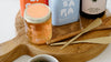 A jar of honey, tins, and a bottle of olive oil are arranged on a wooden cutting board with two brass spreaders.