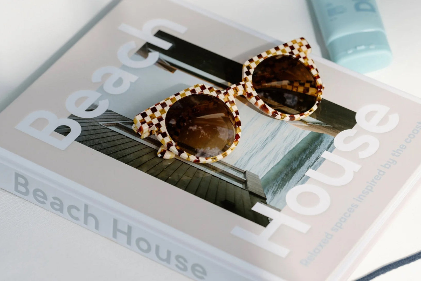A pair of checkered sunglasses rests on a Beach House magazine with a beach house photo on the cover; a blue container is partially visible in the background.