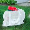 A white mesh tote bag labeled Sunday sits on grass, filled with folded beach towels, with some greenery and rocks visible in the background.