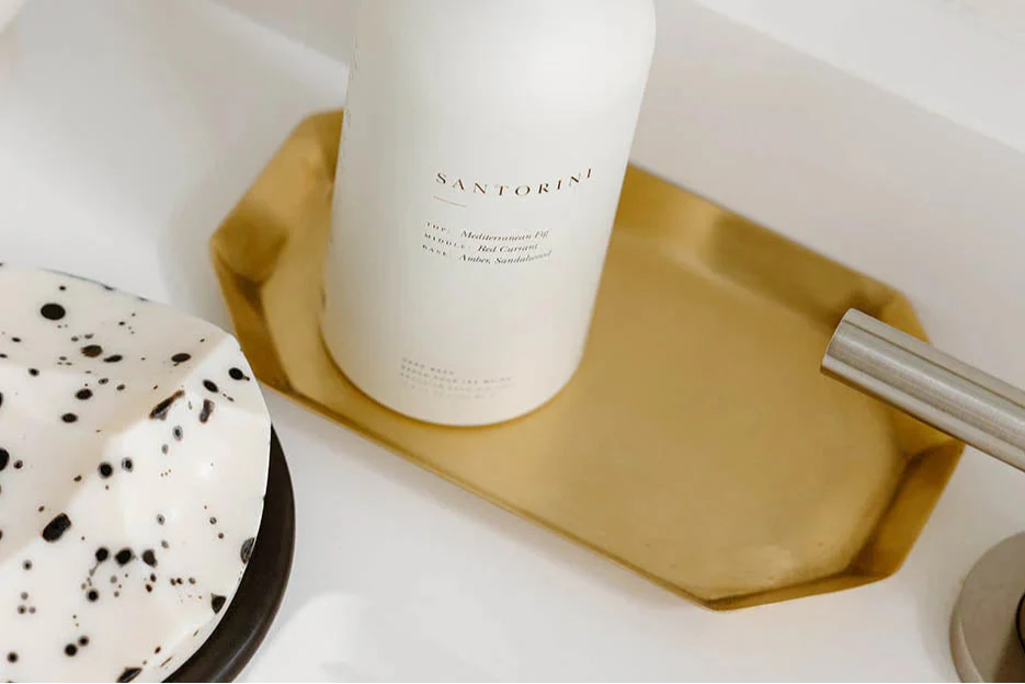 A white bottle labeled Santorini sits on a gold tray next to a white and black speckled dish and a metallic faucet.