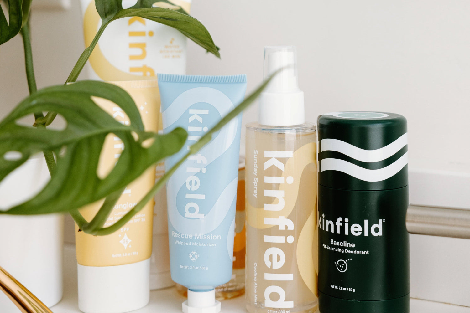 A collection of Kinfield skincare and deodorant products arranged on a bathroom counter beside a green plant.