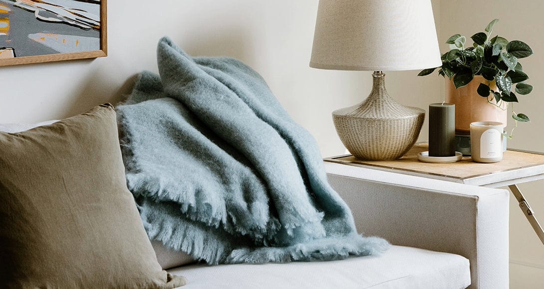 A light blue fuzzy blanket is draped over a white sofa next to a beige pillow, with a lamp, plant, and candles on a wooden side table.