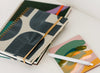 Three notebooks with abstract covers, one secured with an elastic band, and a white pen rest on a white surface.