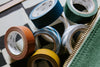 Six rolls of decorative washi tape in various colors and patterns arranged on a checkered surface next to a green zippered pouch.