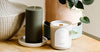 A green ribbed pillar candle and a white scented candle sit on a wooden surface next to ceramic plant pots and a wicker basket.