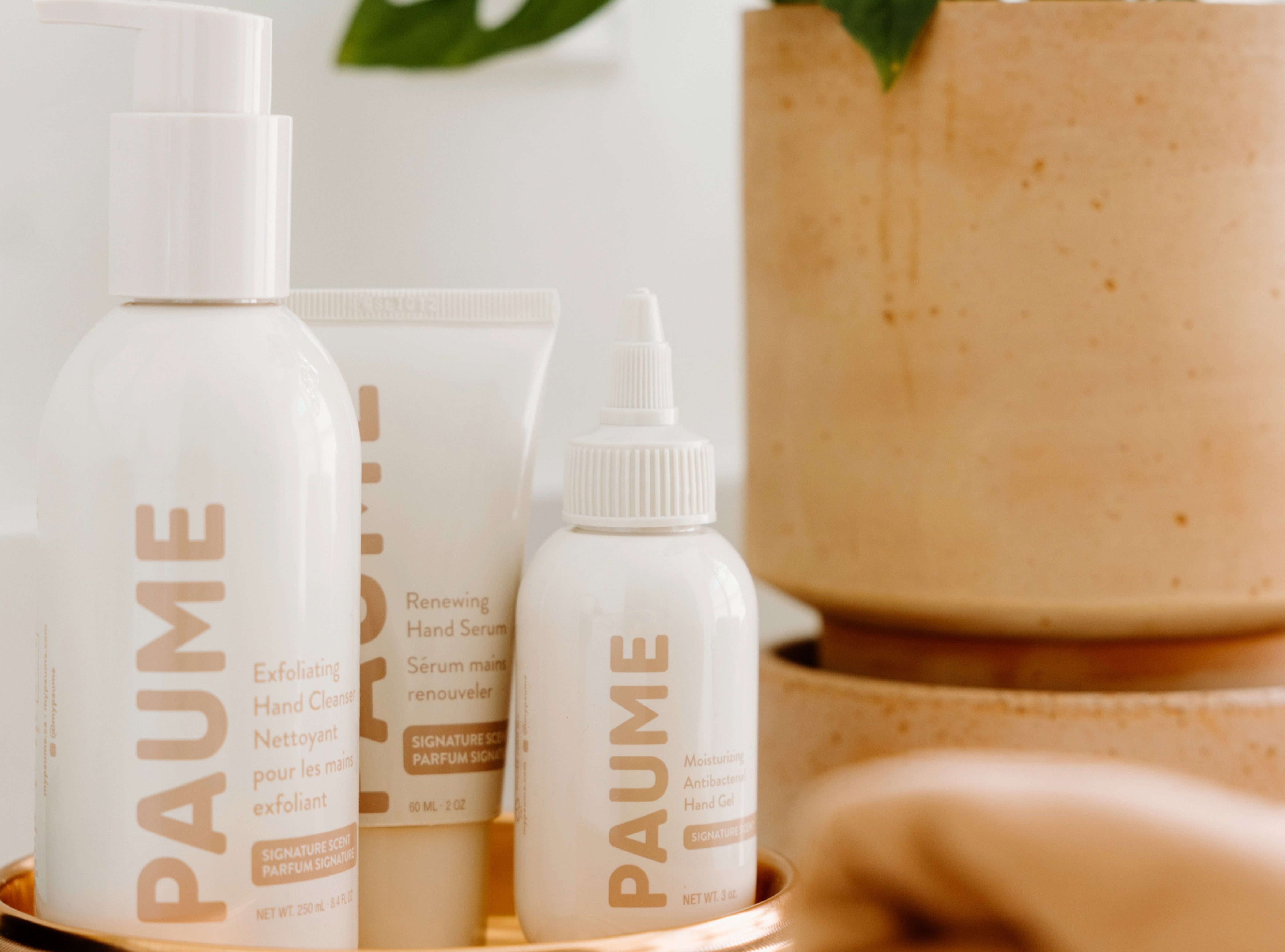 Three PAUME skincare products, including Exfoliating Hand Cleanser, Renewing Hand Serum, and Moisturizing Antibacterial Hand Gel, displayed on a tray beside a tan ceramic pot.