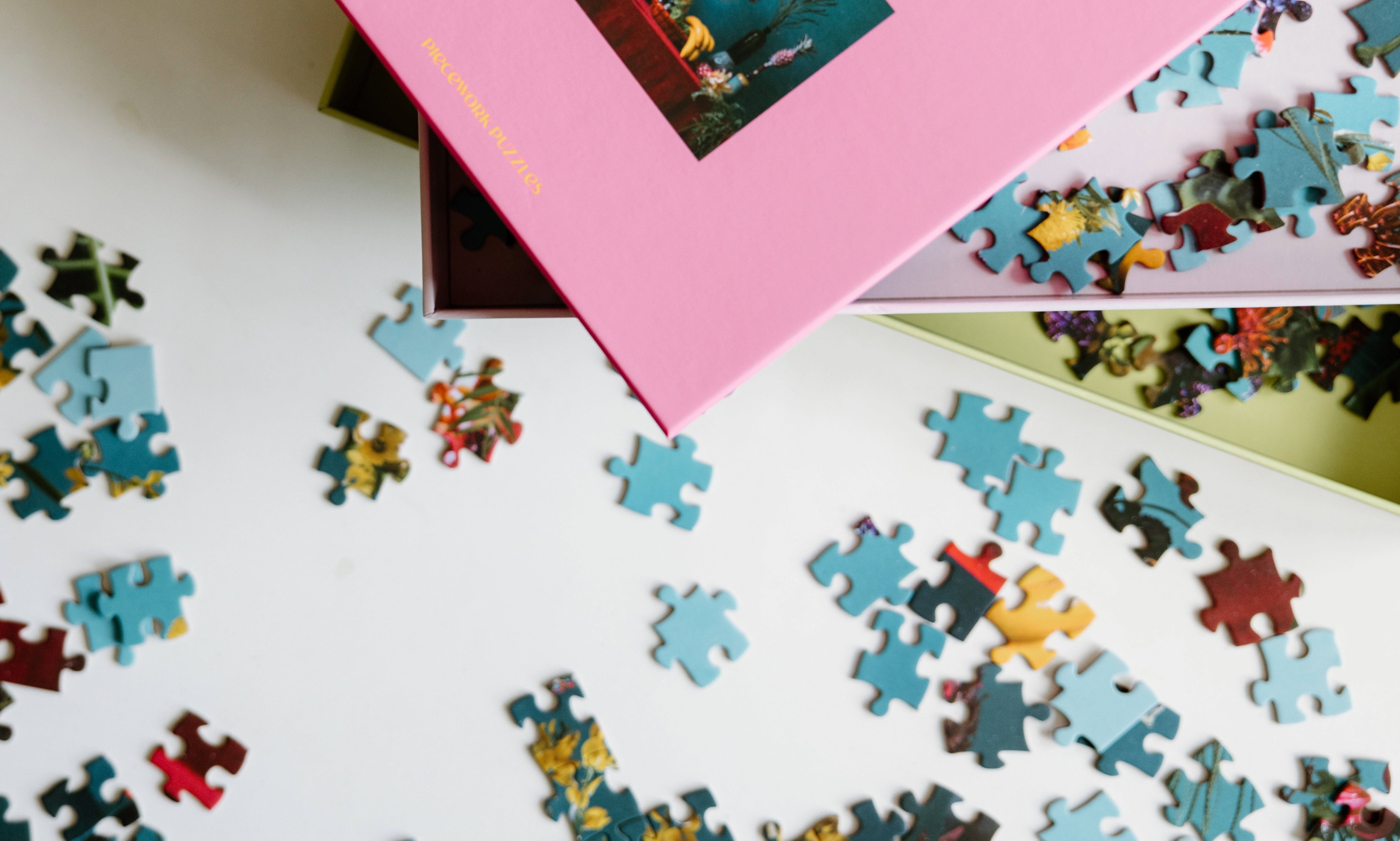 Scattered jigsaw puzzle pieces on a white surface with two partially open puzzle boxes, one pink and one green, visible in the corner.