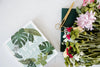A book titled LEAF SUPPLY, gold scissors, a green notebook, and a bouquet of assorted flowers on a white surface.