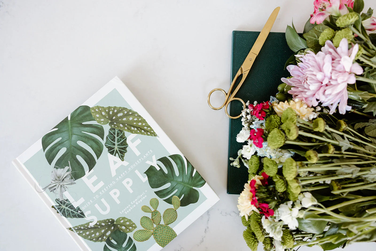 A book titled LEAF SUPPLY, gold scissors, a green notebook, and a bouquet of assorted flowers on a white surface.