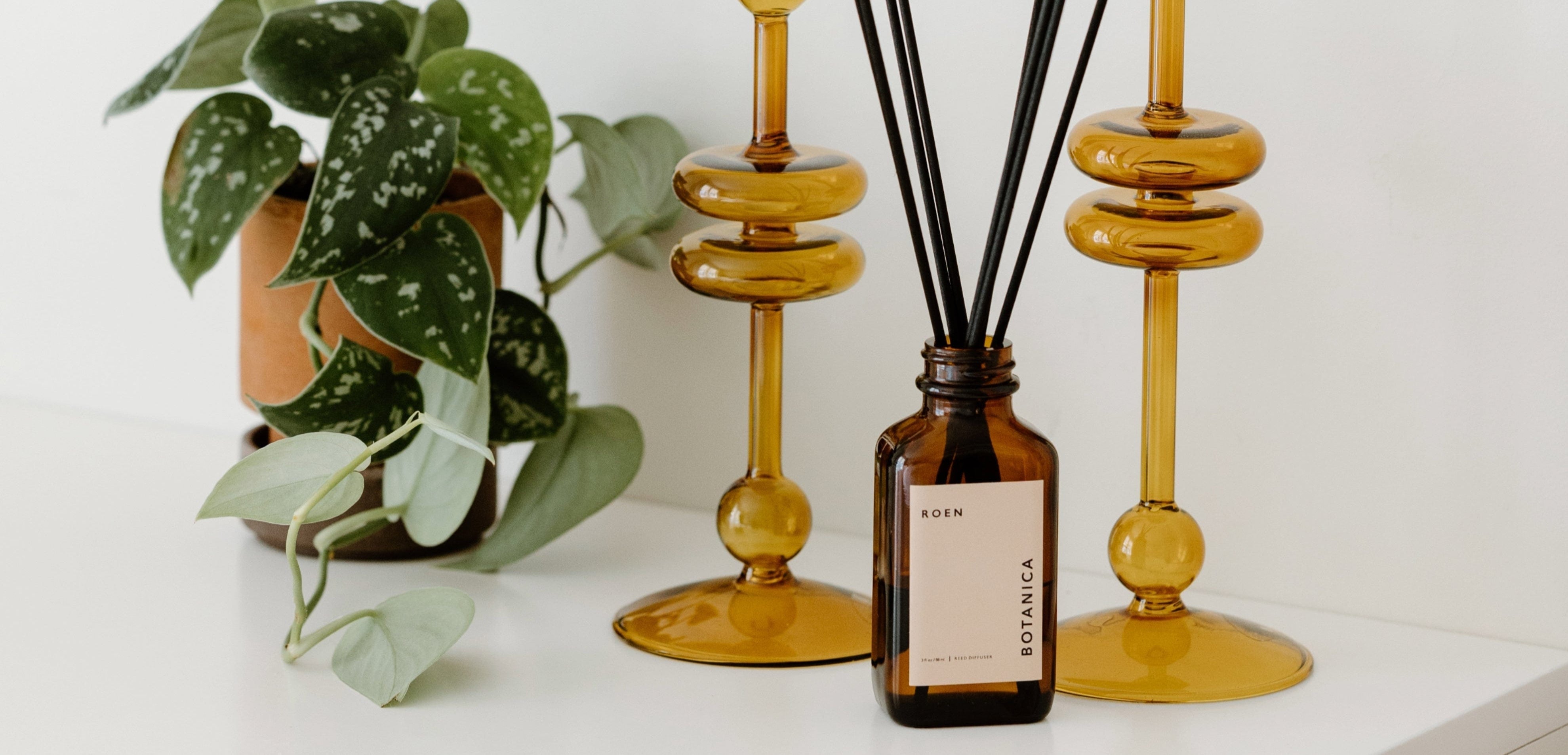 A brown glass diffuser bottle with black reeds sits on a white surface beside two amber glass candleholders and a potted green plant.