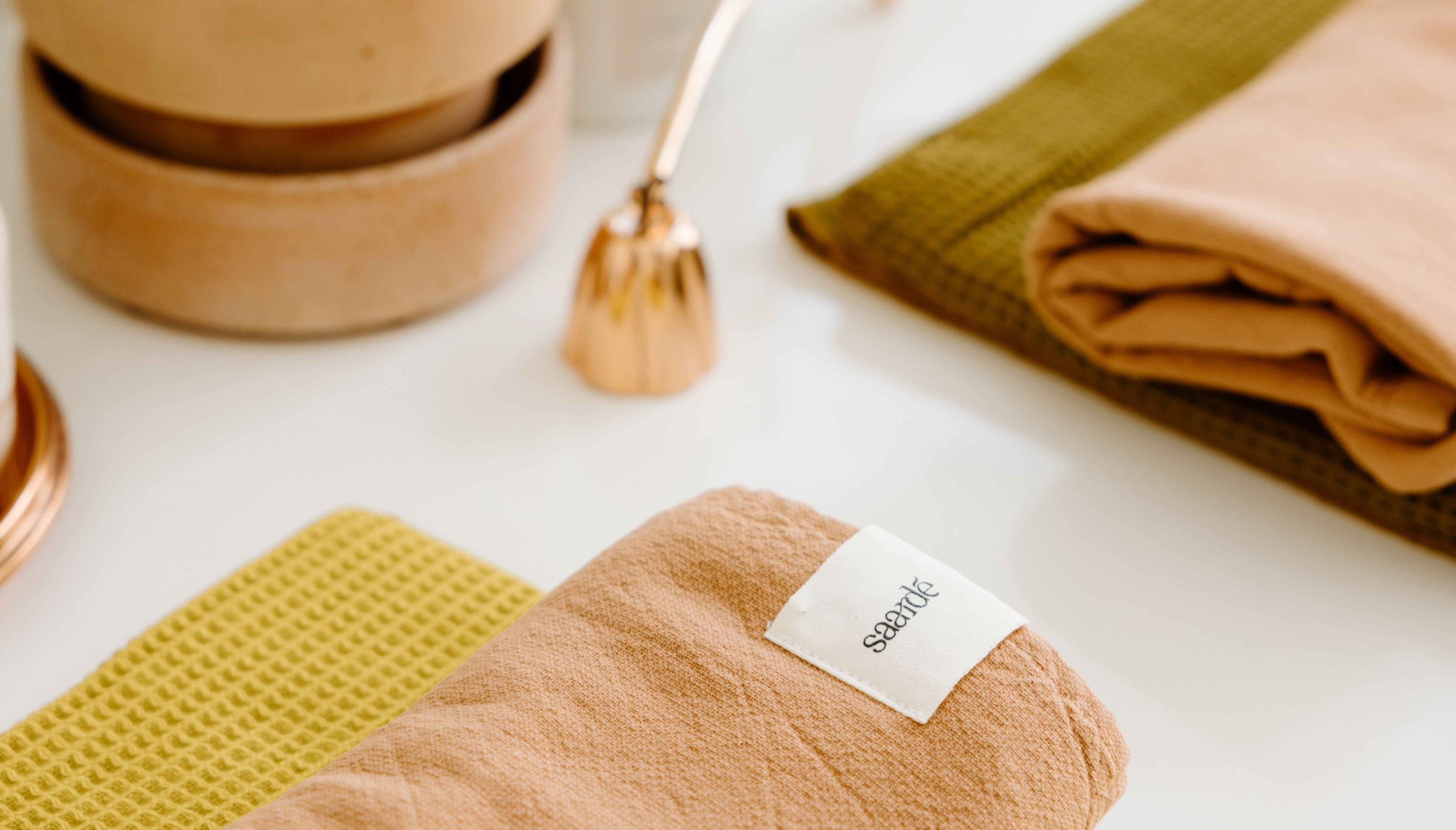 Folded peach and green towels on a white surface with a small gold bell and beige containers in the background. A visible label on one towel reads seadate.