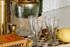 Four clear glasses, a large water dispenser, a jigger, a boxed whiskey set, a lemon, and a checkered tray are arranged on a countertop with a patterned backsplash.