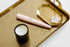 A gold tray holds a white candle in a dark glass jar, a pink electronic lighter, and a burning incense stick on a small gold dish with a white holder.