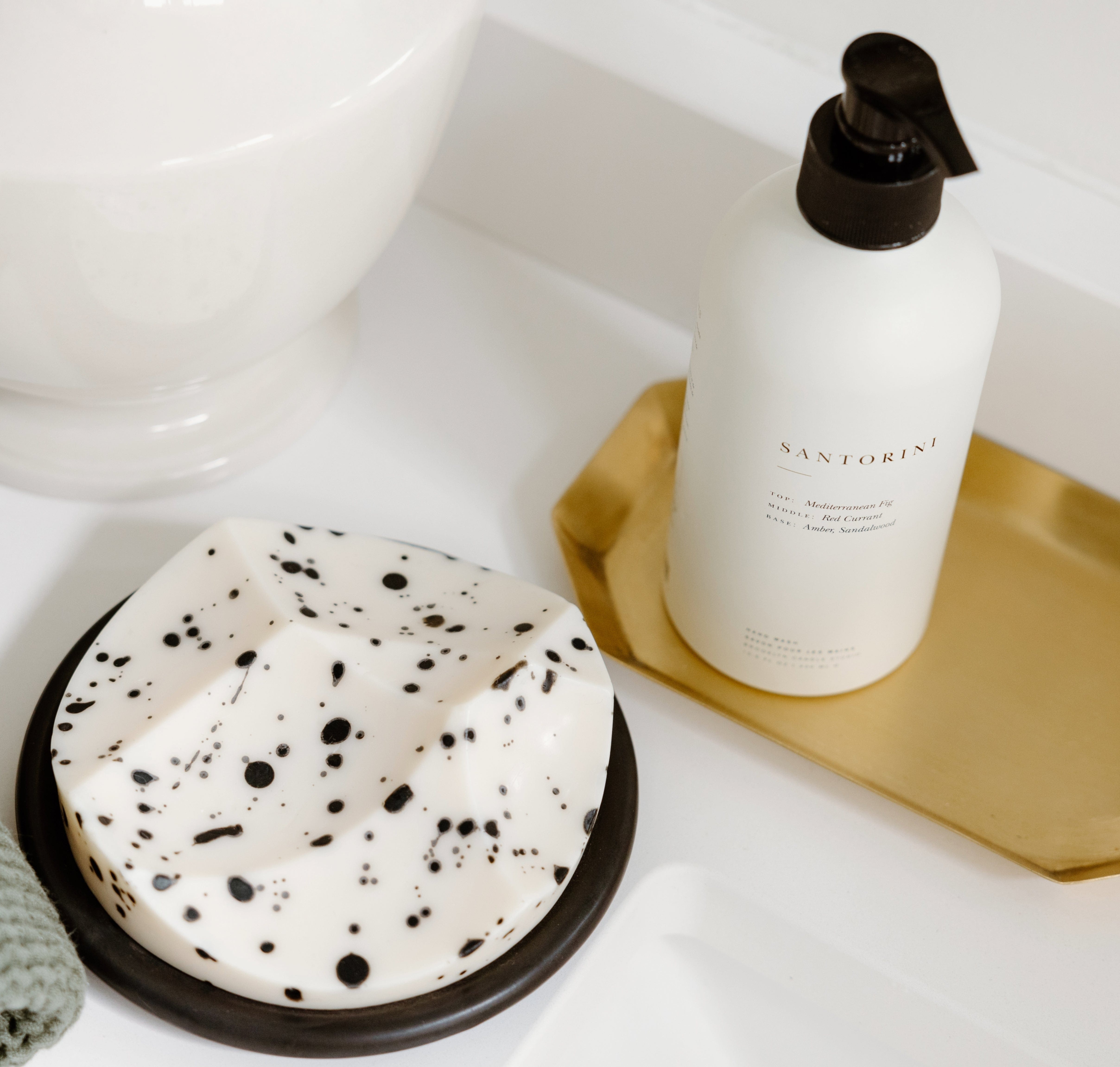 A white bottle labeled Santorini sits on a gold tray next to a white and black speckled soap dish on a bathroom counter.