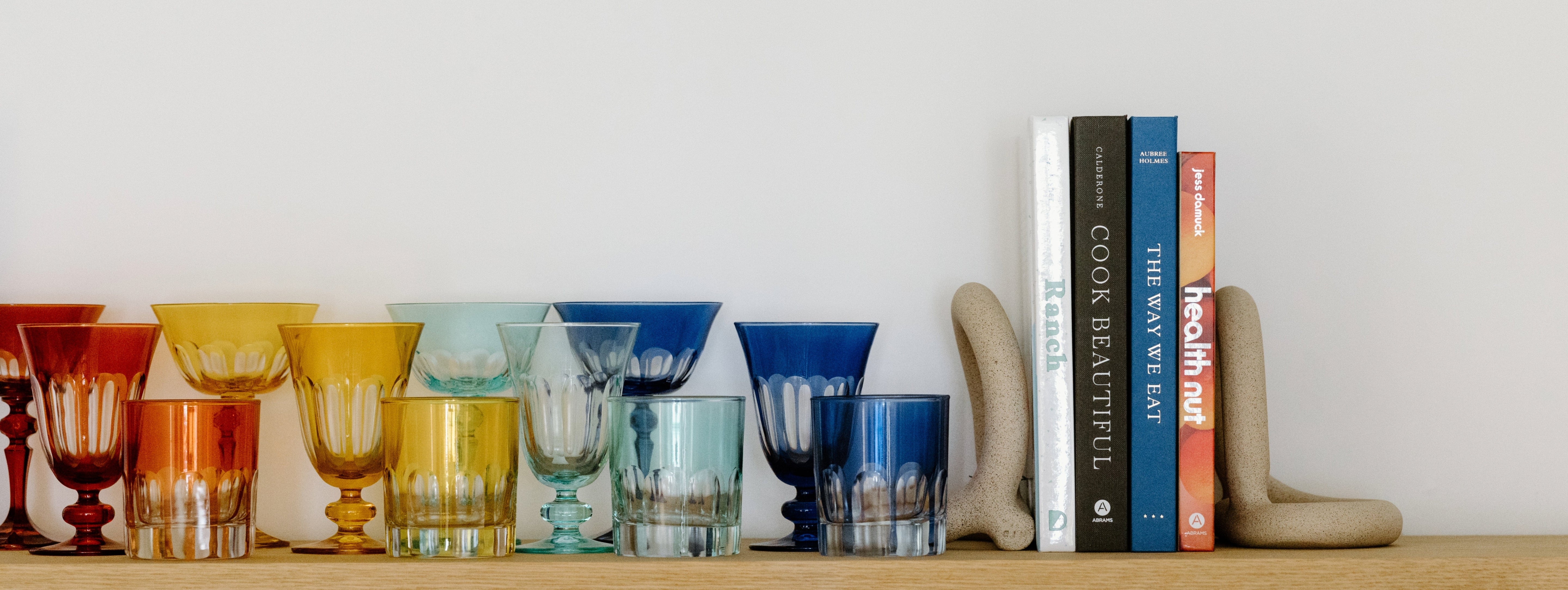 A row of colorful glass cups is arranged by shade next to a set of books held upright by beige bookends on a wooden shelf.