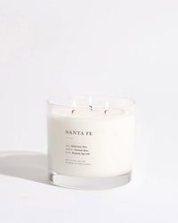 The Brooklyn Candle Studio Santa Fe XL 3-Wick Candle sits on a white surface, its wicks aglow above soy wax. The label highlights earthy notes of orchid cactus, palm, oud wood, sage, and cedar.