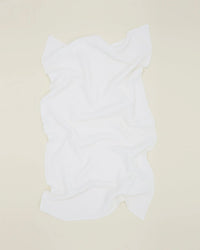 The Hawkins New York Simple Waffle Bath Towel in White lies flat on a pale beige surface, featuring a soft texture, wavy edges, and visible folds that highlight its slightly irregular shape.
