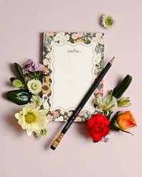 The Flower Market Note Pad by Amy Heitman features floral-patterned tear-off sheets labeled “notes.” It sits on a light pink surface, surrounded by colorful flowers and leaves, with a black pencil with a gold eraser placed diagonally across it.