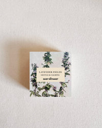 A small box of Lavender Fields - Bar Soap by The Quiet Botanist, handmade in New York and featuring a botanical lavender illustration, rests on a textured off-white surface.