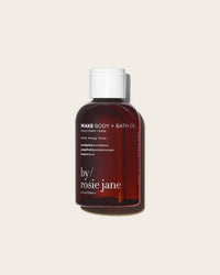 A brown bottle of Rosie Jane Wake Body + Bath Oil with a white cap sits against a beige background. Its label highlights scents of eucalyptus, grapefruit, lemon, and nourishing kakadu plum.
