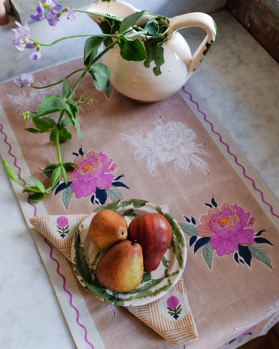 The Babita Table Runner from Soil to Studio’s Valley of Flowers Collection features pink floral patterns, a white pitcher with purple blooms, and a plate of three pears on marble—perfect for setting a beautiful scene.