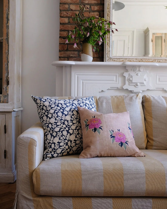 A beige striped sofa with two decorative pillows, including the Soil to Studio Babita Linen Pillow in Old Rose (18x18), sits before a white mantle and exposed brick wall.