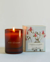 A mini brown glass candle made from natural coconut wax by The Quiet Botanist sits beside its floral Garden Stories: Green Fig & Florals box—a charming travel companion.