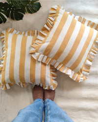 Two Casa Amarosa Golden Yellow Ruffle 16x16in pillows, featuring yellow and white stripes with ruffled edges, rest on beige fabric. A barefoot person in blue jeans stands below, while a green leaf accents the upper left corner.
