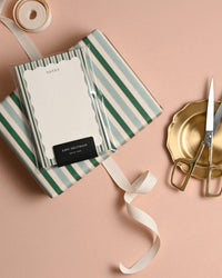 The Amy Heitman Stripes and Scallops Note Pad with tear-off sheets is styled on a pale peach surface with striped wrapping paper, gold scissors, ribbon, and a gold tray. An attachable magnet adds convenience.