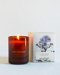 The Quiet Botanist’s In the Weeds - Mini Candle, a natural coconut wax candle in a brown glass jar, sits beside its box decorated with purple-blue flowers and labeled "In the Weeds" and "The Quiet Botanist.