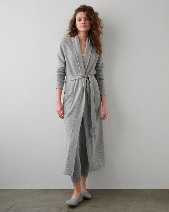 A woman with loose curly hair stands against a light gray wall, wearing the White + Warren Cashmere Long Robe in Grey Heather, matching pants, and gray slippers. She looks relaxed and casual.