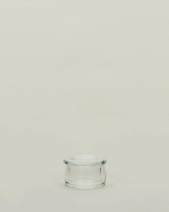 A small, empty Simple Glass Butter Keeper by Hawkins New York with a lid sits centered on a plain, light beige background, evoking minimalist style and leaving ample empty space above.