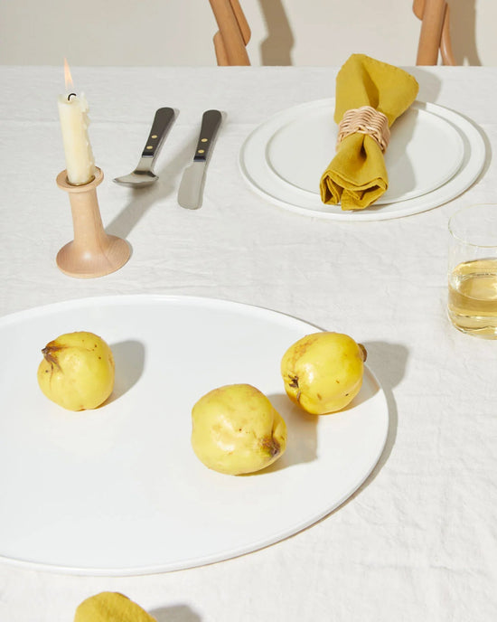 A minimal table setting with a white tablecloth, Hawkins New York’s Simple Porcelain Serving Platter holding three yellow quinces, a drink, candle, cutlery, and a plate with a mustard yellow napkin—all in bright lighting.