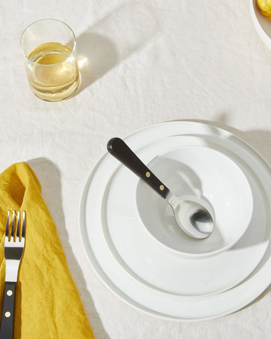 A table setting with a Hawkins New York Simple Porcelain Dinner Plate, matching bowl, a spoon in the bowl, a fork on a yellow napkin, and a glass of light-colored drink atop a crisp white tablecloth.